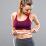 Fitness Woman Measuring Her Body Isolated On Gray Background. Weight Loss Concepts